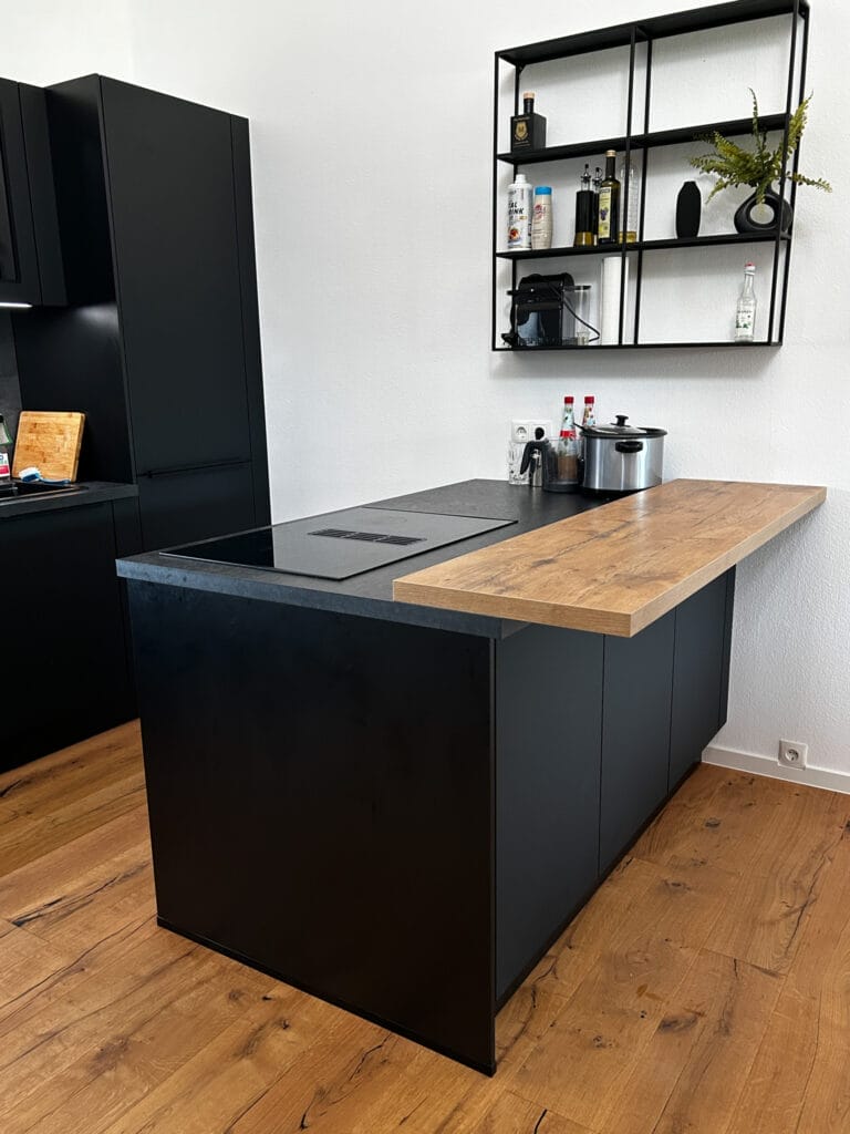 Schwarze Kochinsel mit integrierter Holzarbeitsfläche und offenen Regal auf Holzparkett.